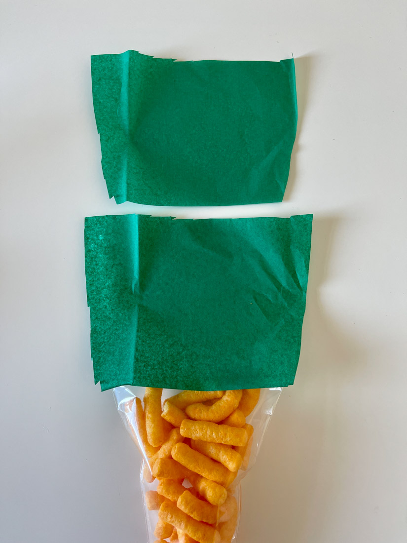 Two sheets of green tissue paper, candy cone filled with cheesy chips on table