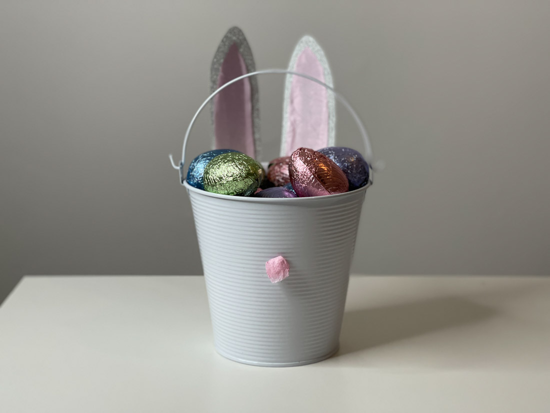 Easter Bunny Bucket Basket with bunny ears and pink tissue nose filled with chocolate eggs sitting on white table.